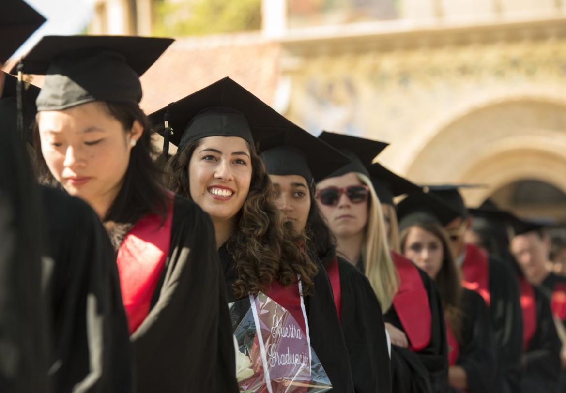 Students in cap and gown lined up under Stanford arches, smiling student in focus